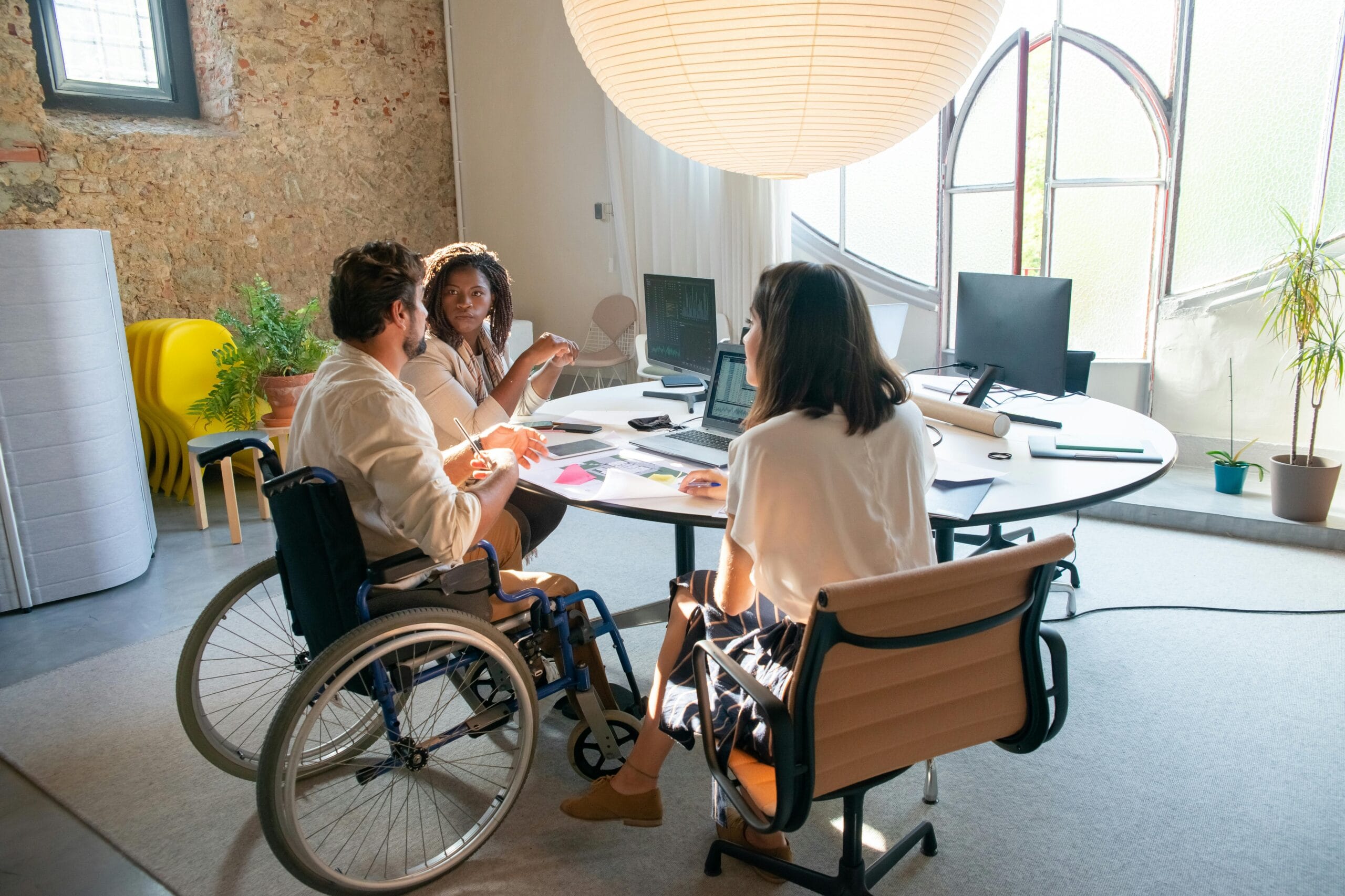 3 people working together at a table with laptop, well lit big room, one is a wheelchair user.